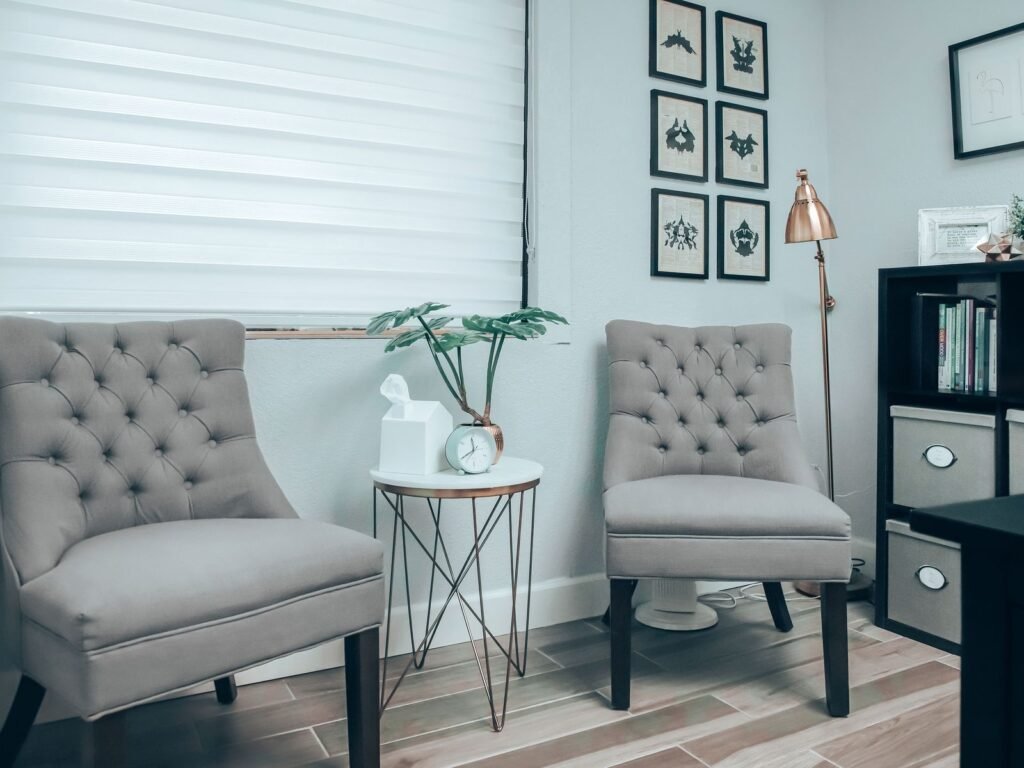 Calm, modern consultation room with two gray tufted chairs, a small side table with a plant and framed prints on the wall