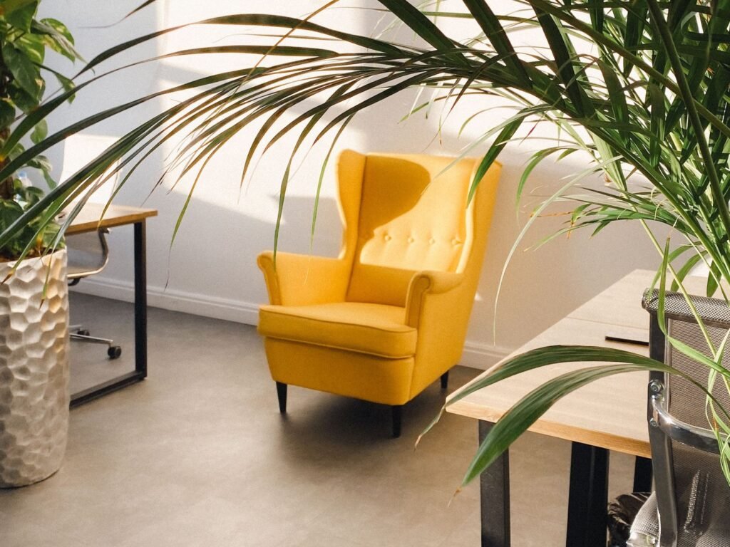 Sunlit room with a yellow wingback armchair, potted palms and a light wood desk